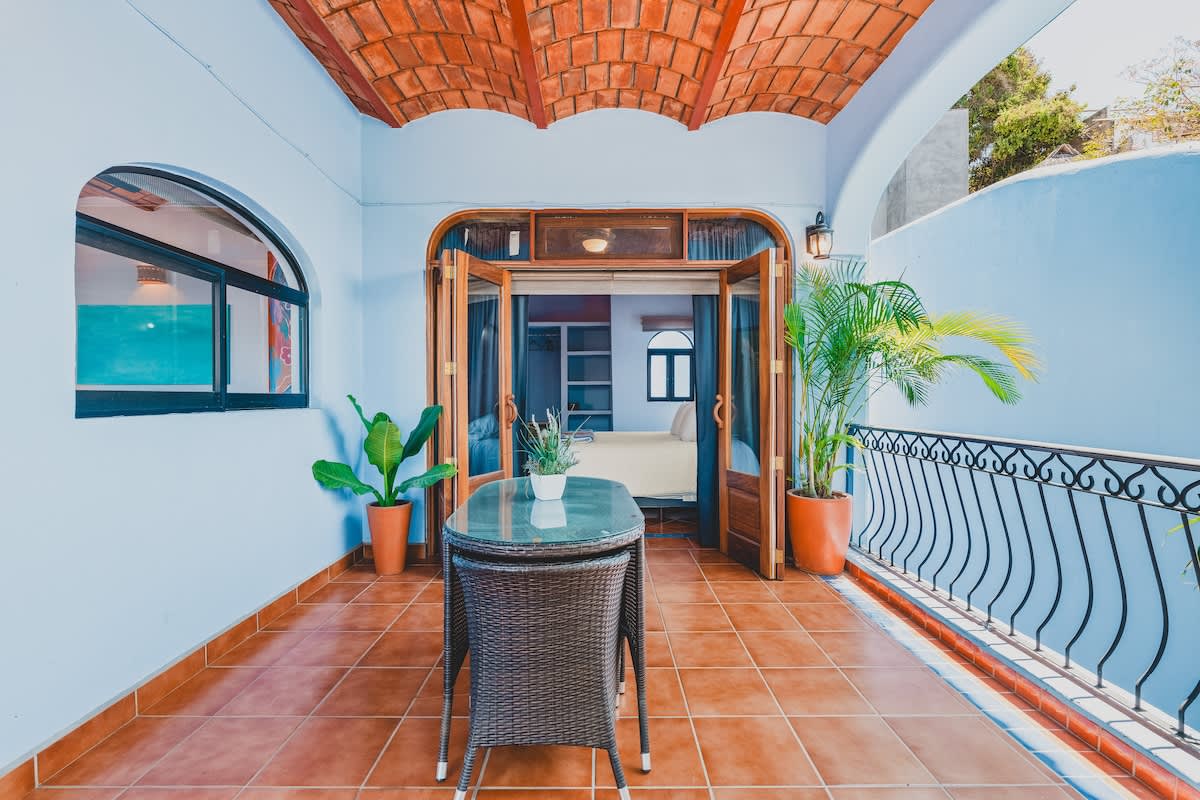 An inviting outdoor balcony features terracotta floor tiles and a terracotta vaulted ceiling, with light blue walls. A glass-top table with a wicker base is centered, and double wooden doors open into a bedroom, flanked by potted plants and an arched window.
