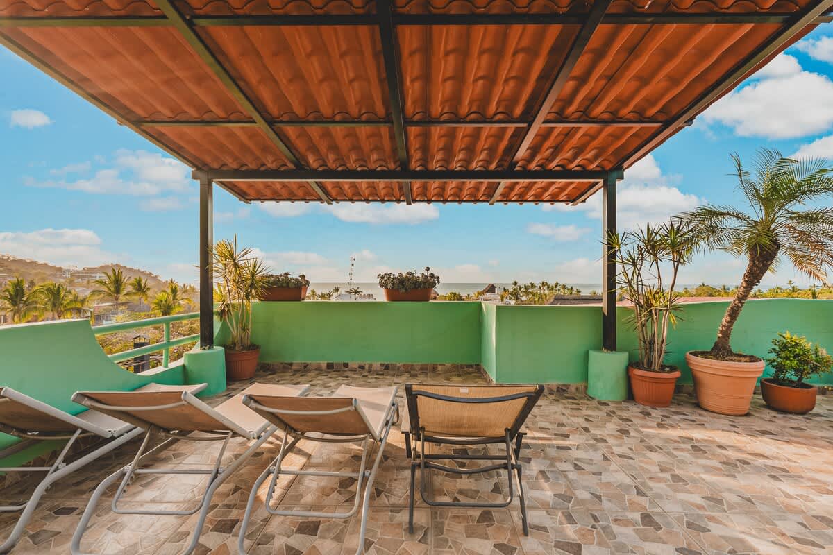 A shaded section of the rooftop terrace features several lounge chairs on patterned tile, facing a green wall and offering a view of the distant ocean and surrounding town under a bright sky.