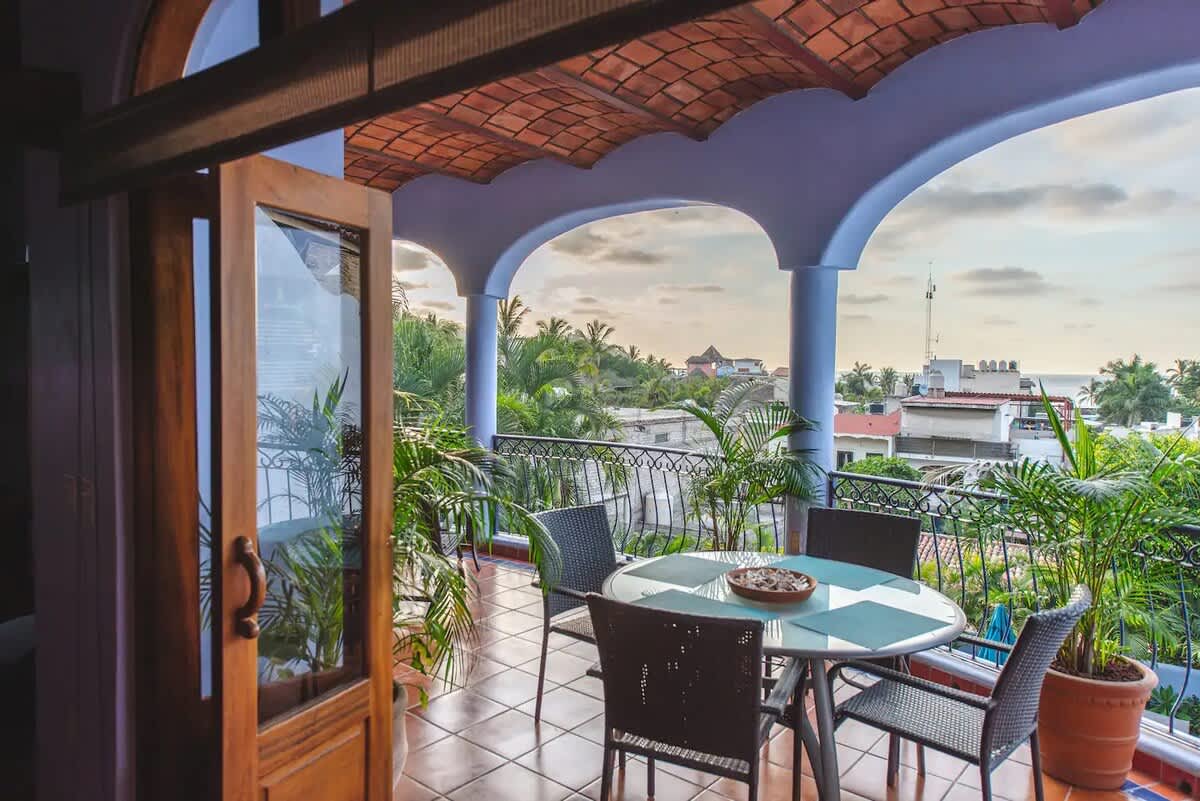 Spacious balcony with a glass-top dining table and chairs, offering panoramic views of the town, palm trees, and distant ocean under a cloudy sky.