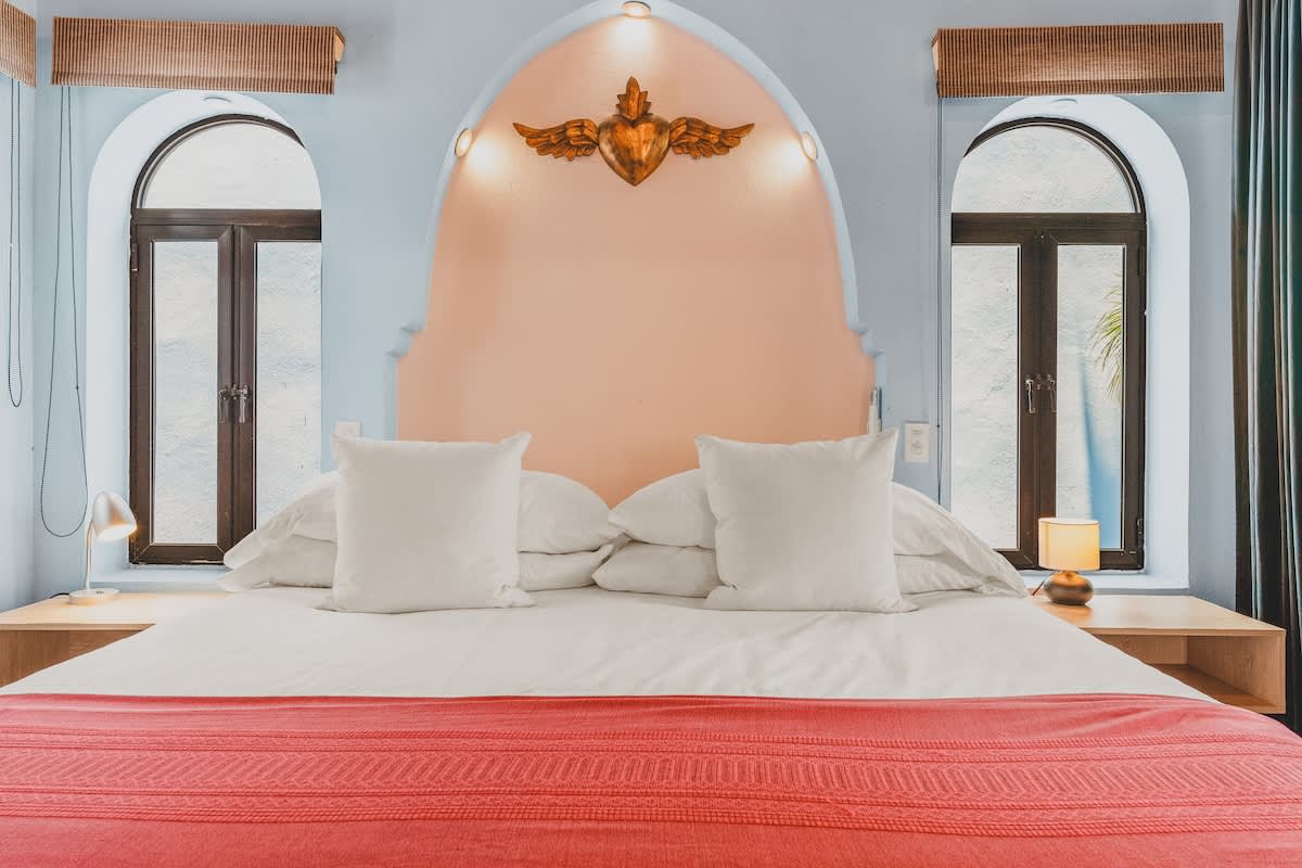 A close-up of a bedroom shows a large bed with a white duvet and red throw blanket, flanked by two light wood nightstands with lamps. The bed is set against a light pink arched headboard with a golden winged ornament, with arched windows on either side.