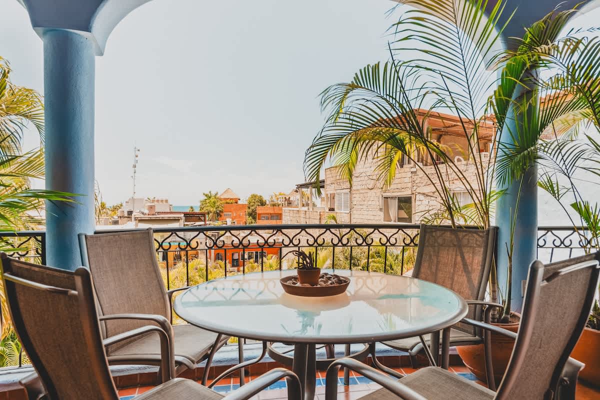 A spacious balcony featuring a round glass-top table with four chairs, surrounded by potted palm trees and offering a panoramic view of the urban landscape.