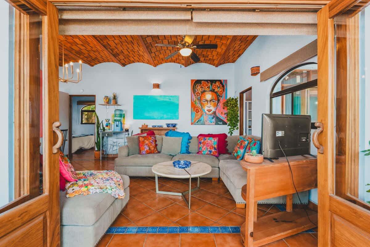 A wide shot of the living area from the balcony entrance, showcasing a large sectional sofa with colorful pillows, a coffee table, and vibrant artwork on light blue walls, with a dining area visible beyond.