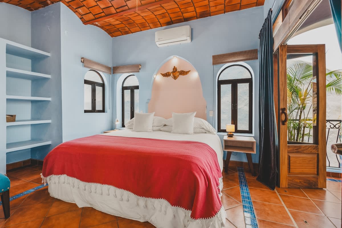 A spacious bedroom features light blue walls, a vaulted terracotta tile ceiling, and terracotta floor tiles with blue decorative accents. A large bed with a white duvet and red throw blanket is centered against a light pink arched headboard with a golden winged ornament.