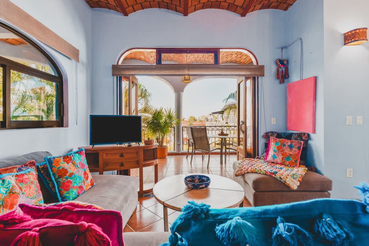 A bright living room with light blue walls, a large sofa with colorful cushions and throws, a wooden coffee table, and a TV, with an open view to a sunny balcony.