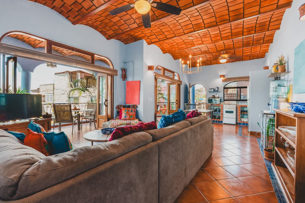 A vibrant living room with light blue walls, a vaulted terracotta tile ceiling, and a large sectional sofa adorned with colorful pillows, opening to an outdoor balcony.