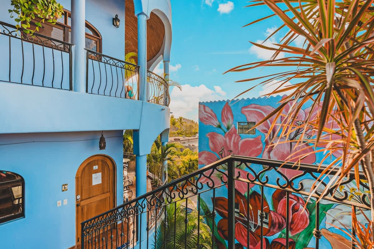 The vibrant exterior of the property showcases light blue walls, arched windows, and balconies with ornate black railings, with a colorful floral mural on an adjacent building and lush tropical foliage.