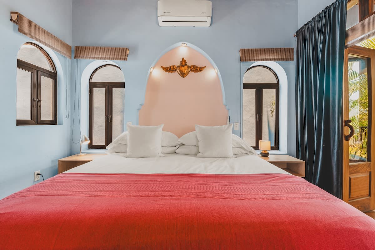A bright bedroom features light blue walls and a terracotta vaulted ceiling, with a large bed dressed in white and a red throw blanket. An arched light pink headboard with a golden winged ornament is centered between two arched windows, and an air conditioning unit is mounted above.