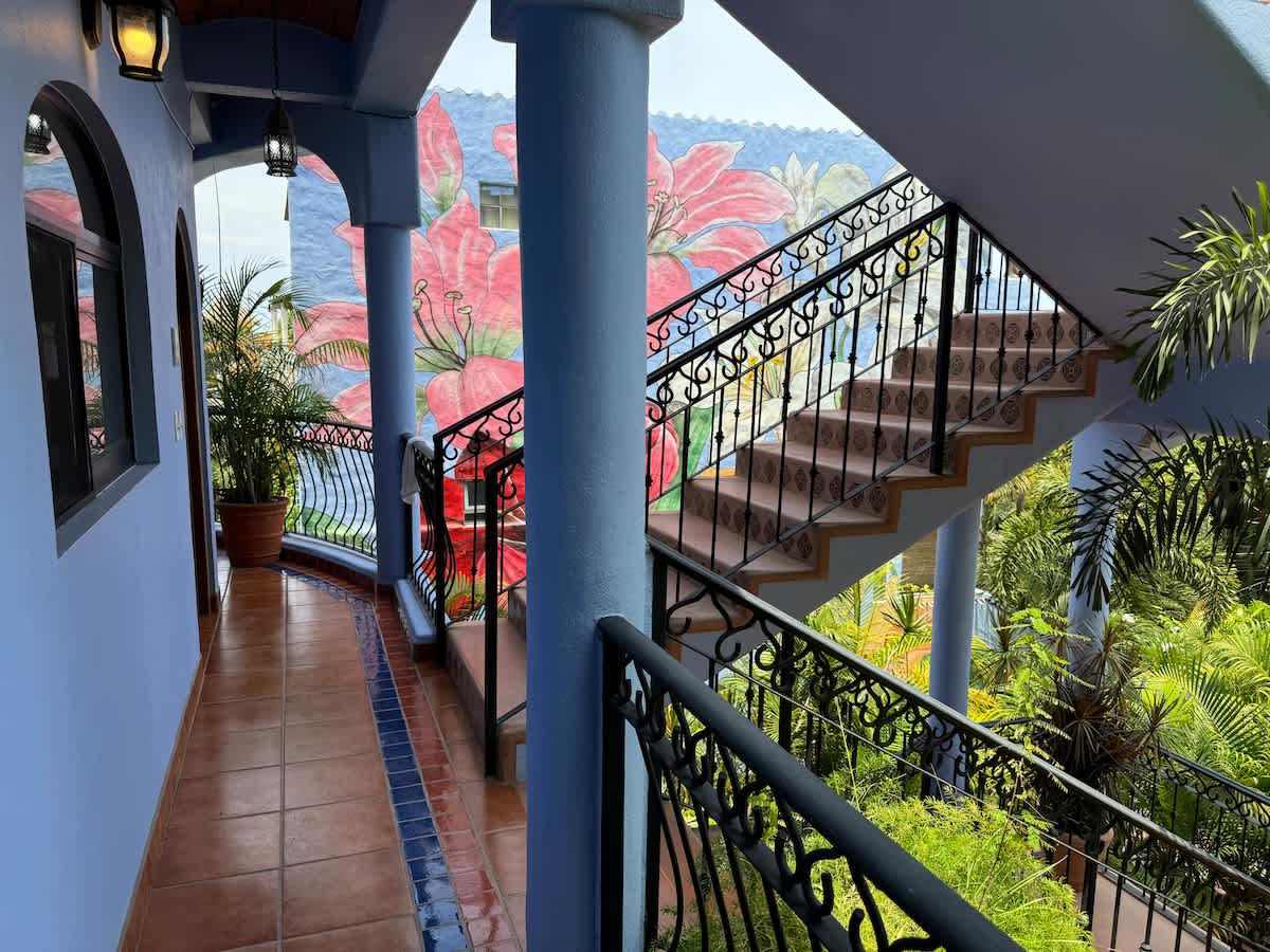 Open-air hallway with light blue walls, terracotta tiled flooring, and a black wrought iron railing, leading to a staircase with a vibrant floral mural on the wall.