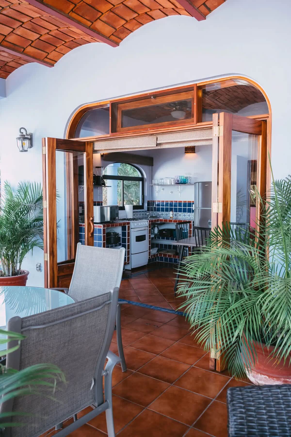 Outdoor patio with terracotta tile flooring and a glass-top dining table, looking into a kitchen with blue and white tiled walls and wooden arched doors.