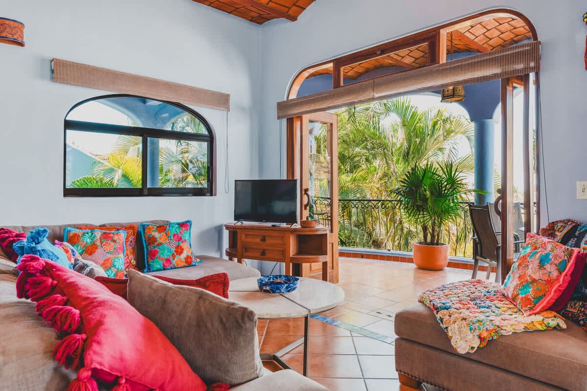 A cozy living room featuring a TV on a wooden stand, a comfortable sofa with bright floral pillows, and large glass doors leading to a lush green balcony with palm trees.