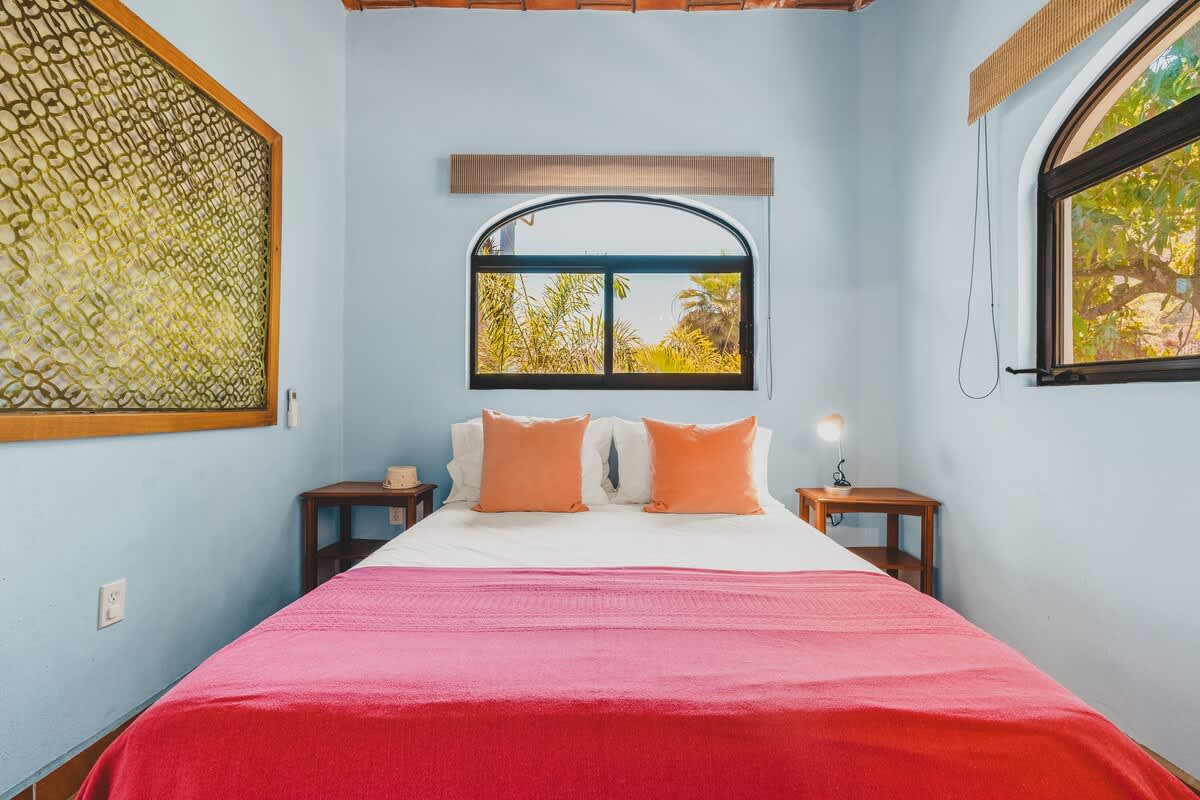 A cozy bedroom features light blue walls, a queen-sized bed with white linens, orange pillows, and a bright pink blanket, set against a terracotta tile floor with a decorative border and an arched window overlooking lush greenery.