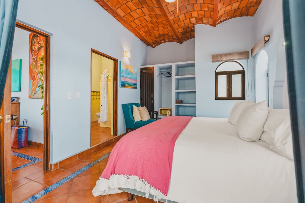 Cozy bedroom with a large bed, light blue walls, and a vaulted terracotta tile ceiling, showing an open doorway to a bathroom and built-in shelving, with colorful artwork on the walls.