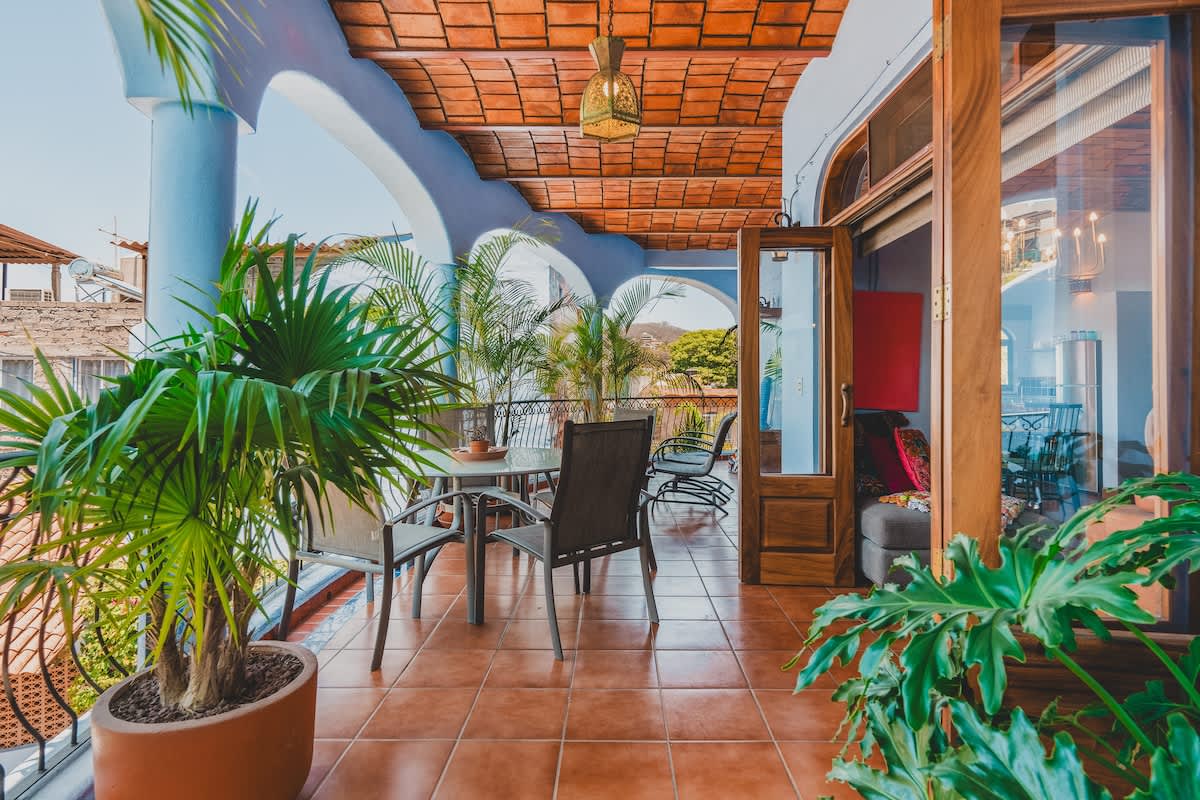 A sunny balcony with terracotta tile flooring, featuring an outdoor dining set, potted palm trees, and arched architectural details, overlooking a tropical landscape.
