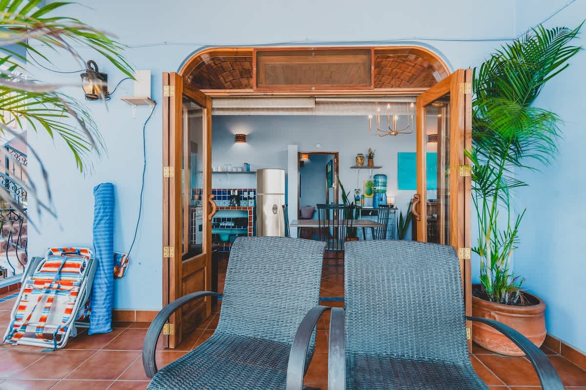 An outdoor balcony with two dark wicker chairs, looking through open wooden doors into the bright kitchen and dining area of the apartment.