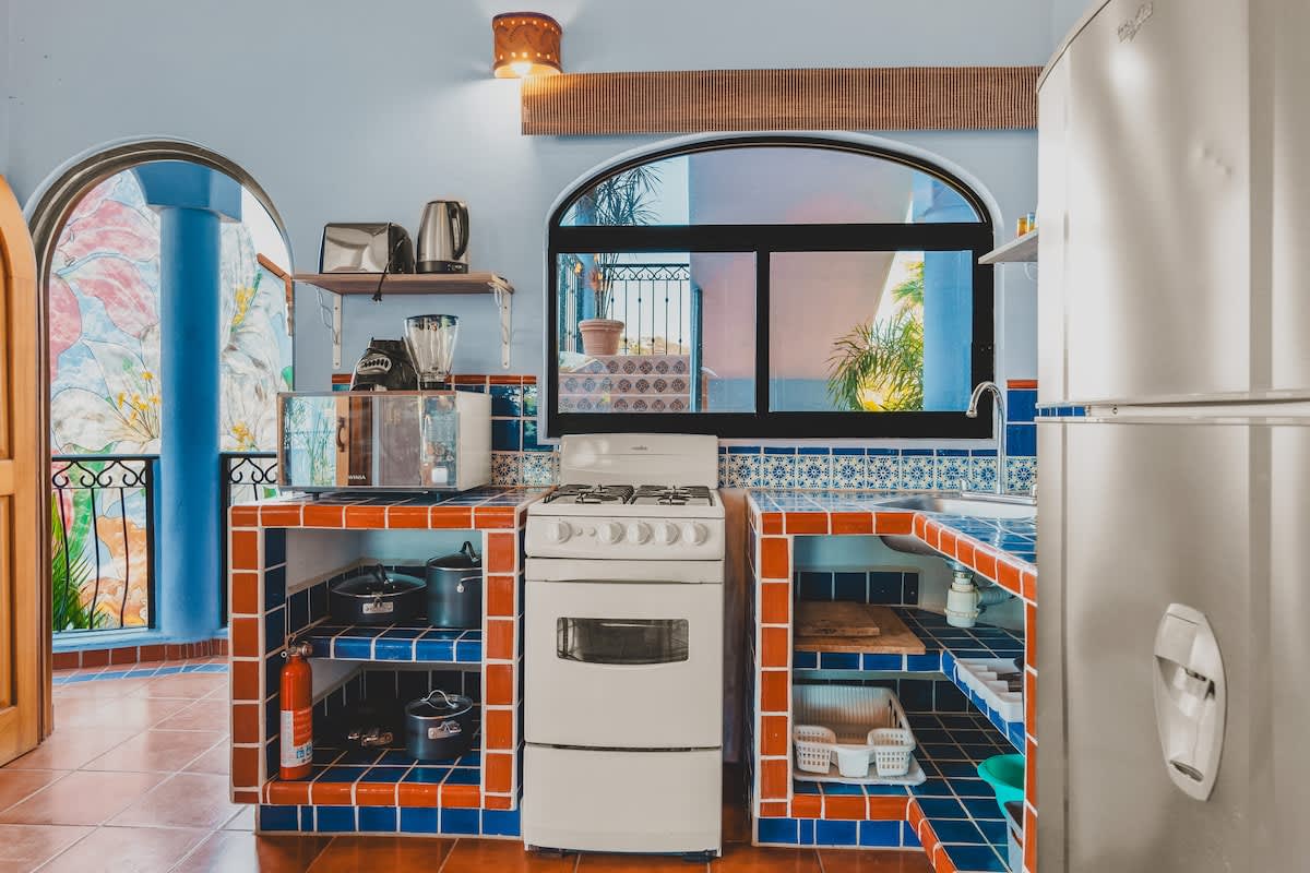 A well-equipped kitchen with a white gas stove, blue and orange tiled countertops and open shelving, a microwave, and an arched window, showcasing a vibrant, functional space.