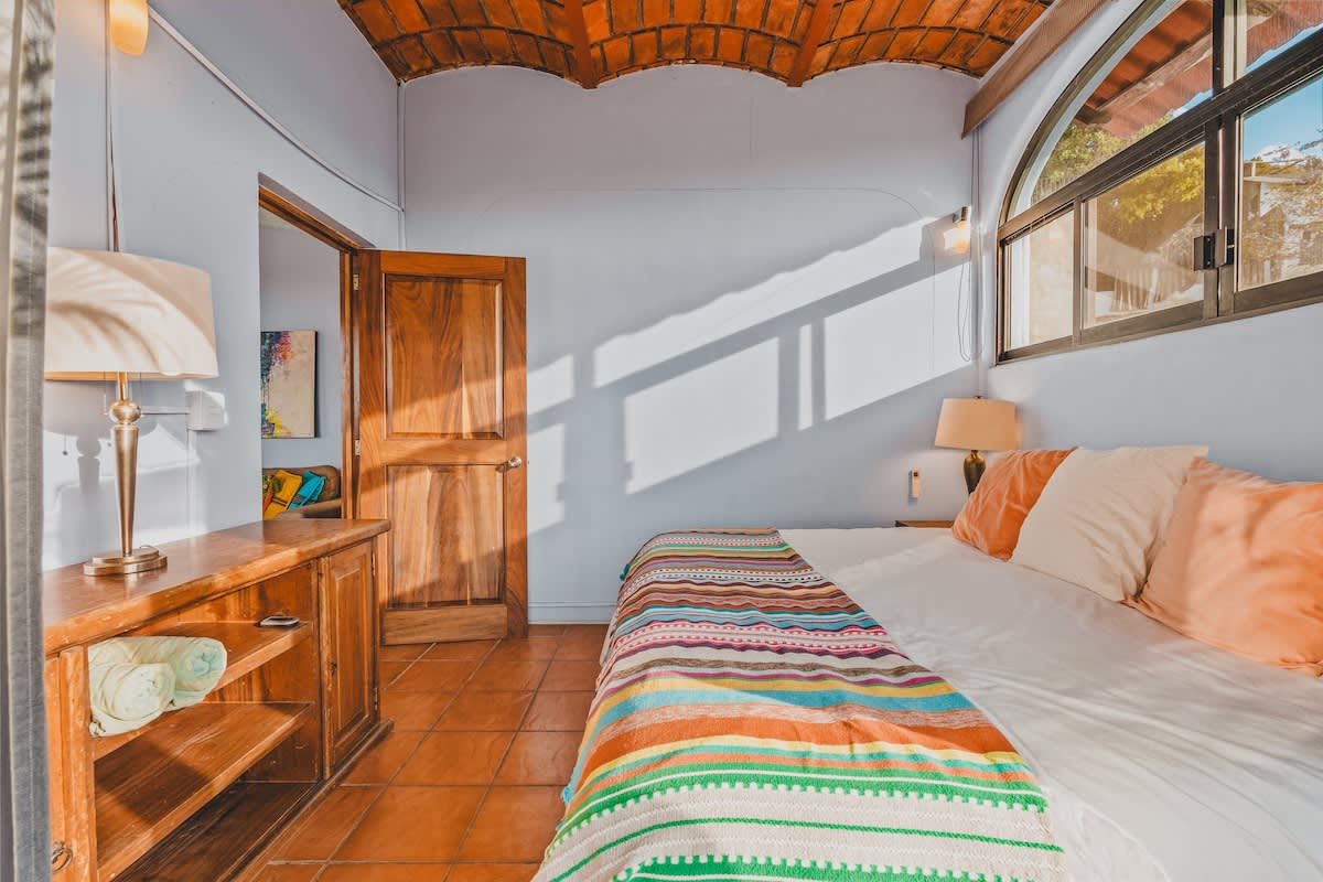 Another view of the bedroom, showing a wooden nightstand with a lamp, a wooden door, and the bed with a colorful blanket, all under a vaulted terracotta-tiled ceiling.