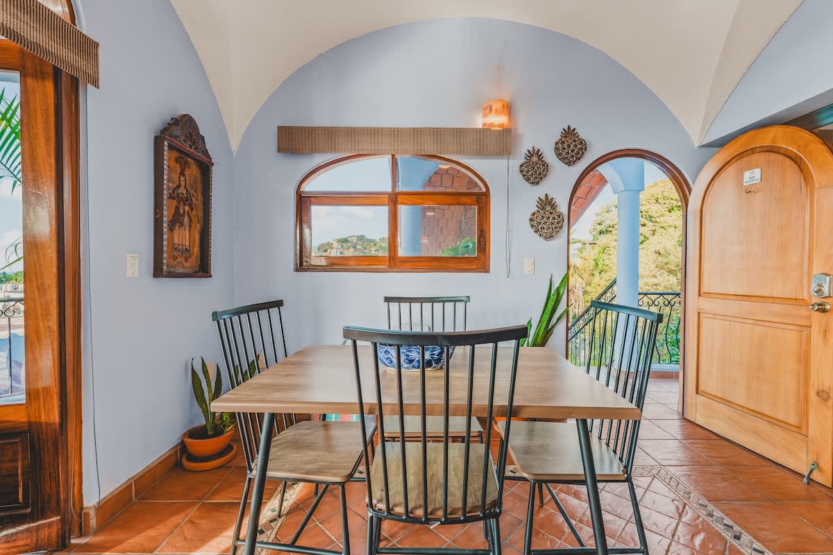 A cozy dining area features a light wood rectangular table with black metal chairs, set on terracotta tile floors with decorative inlays. The room has light blue arched walls, a large arched window, and decorative pineapple wall art.