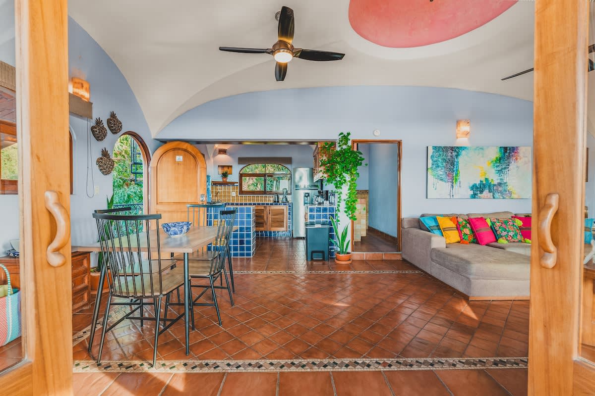 Open-concept living space showing a dining area with a wooden table and black chairs, transitioning into the kitchen with blue and orange tiles, and the living room with a sofa.