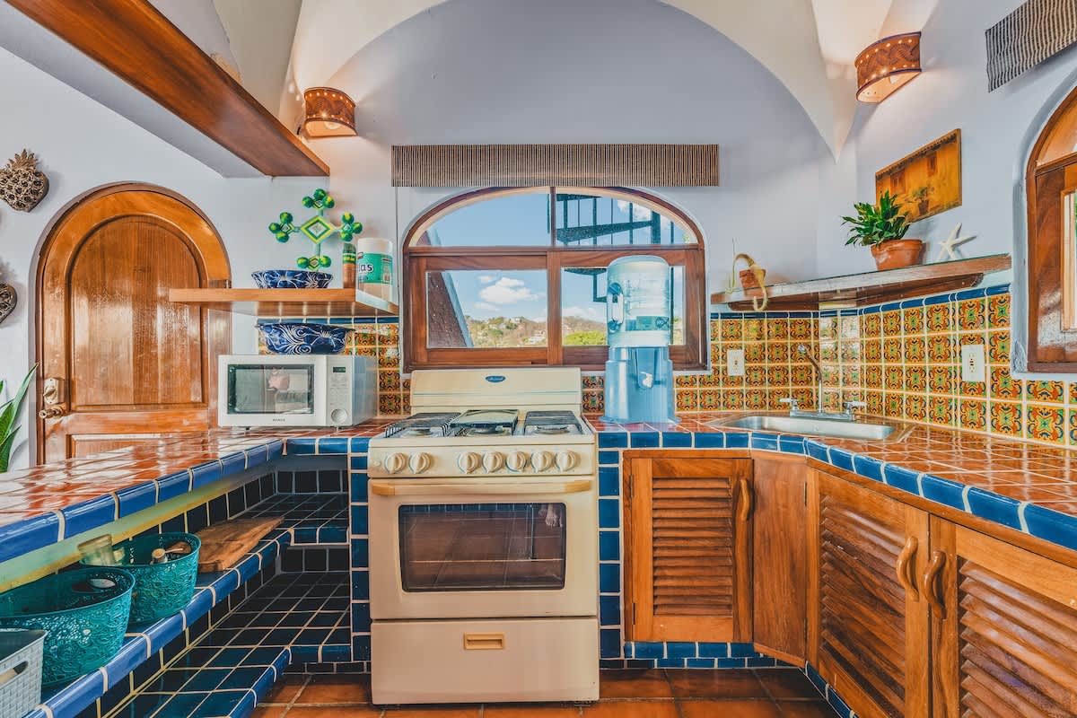 A colorful kitchen with blue and orange tiled countertops, a white gas stove with an oven, a microwave, and a large water dispenser. Arched wooden windows offer a view of the surrounding landscape.