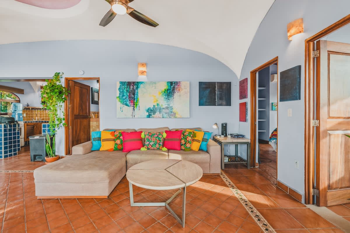 Spacious living room with a light brown sectional sofa adorned with bright, colorful pillows, a round coffee table, and a large abstract painting on the wall, leading to the kitchen area.