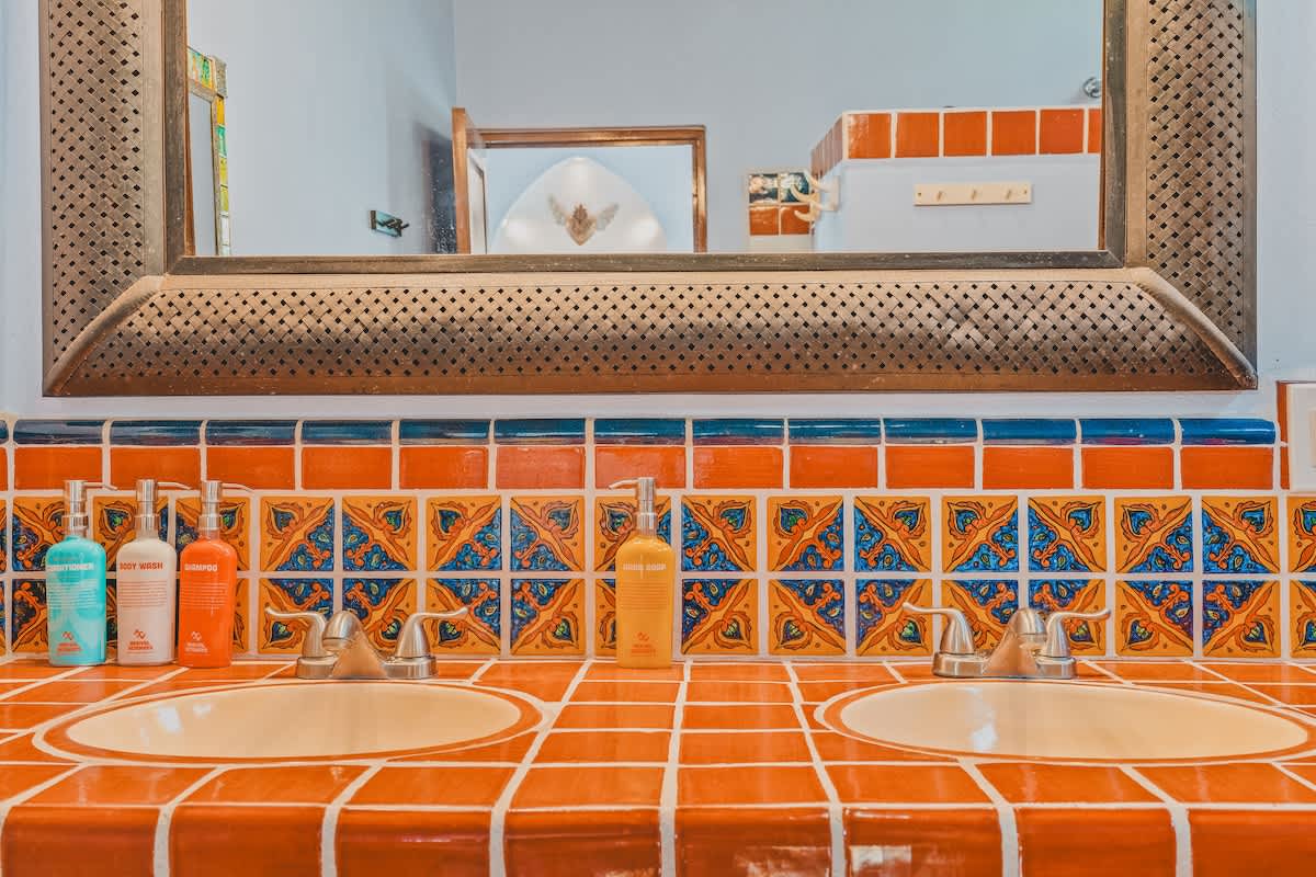 Close-up of the double vanity in the bathroom, showcasing the orange tiled countertop, decorative blue and orange patterned backsplash tiles, two sinks with chrome faucets, and various toiletries.