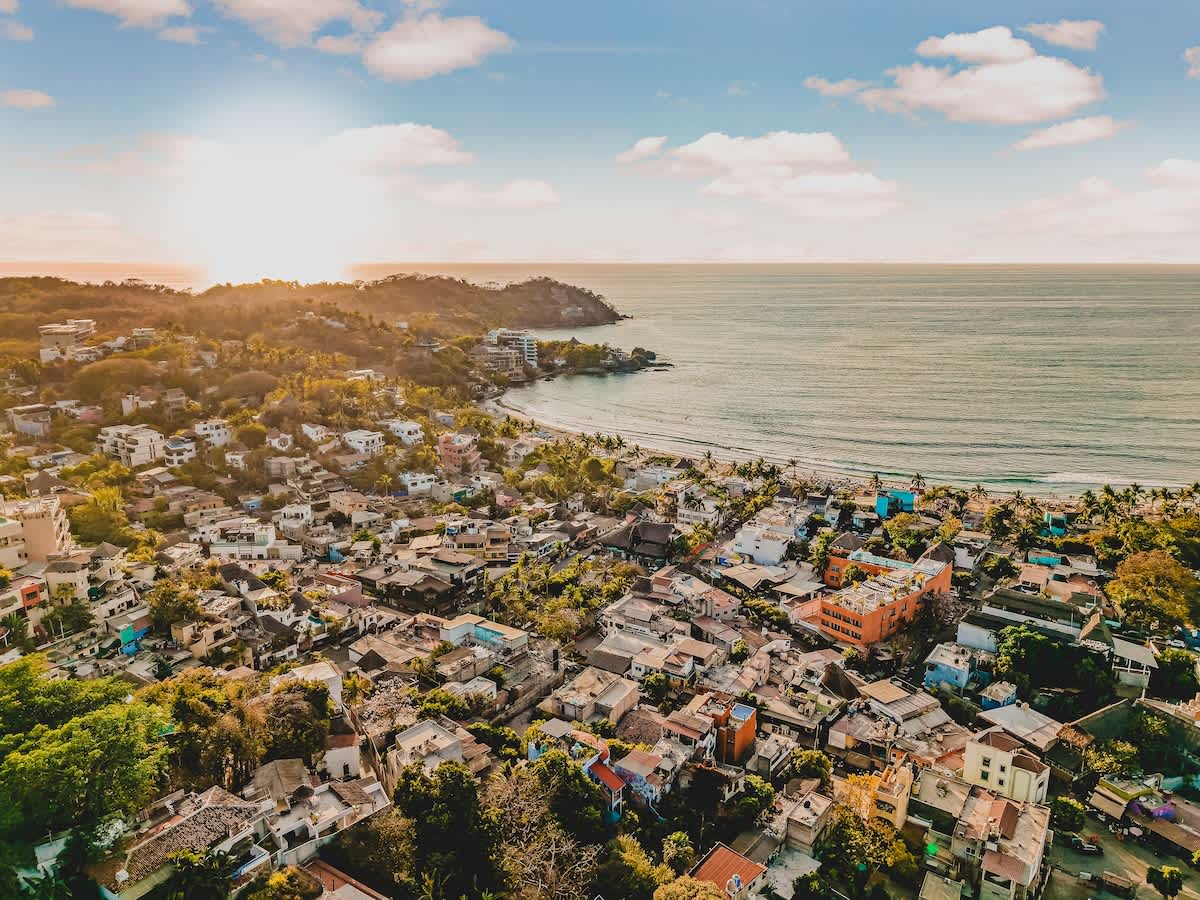Aerial view of a vibrant coastal town with numerous houses nestled among lush green hills, leading down to a sandy beach and the sparkling ocean under a bright, partly cloudy sky. The sun casts a warm glow over the landscape.