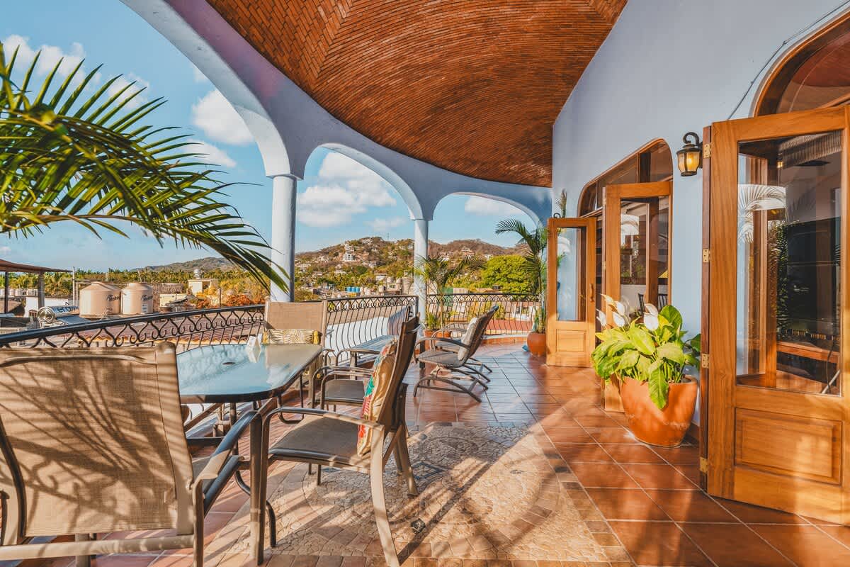 A long, covered balcony with terracotta-tiled flooring and multiple outdoor seating arrangements, including a dining table and lounge chairs. It offers a panoramic view of rolling hills and the vibrant town below.