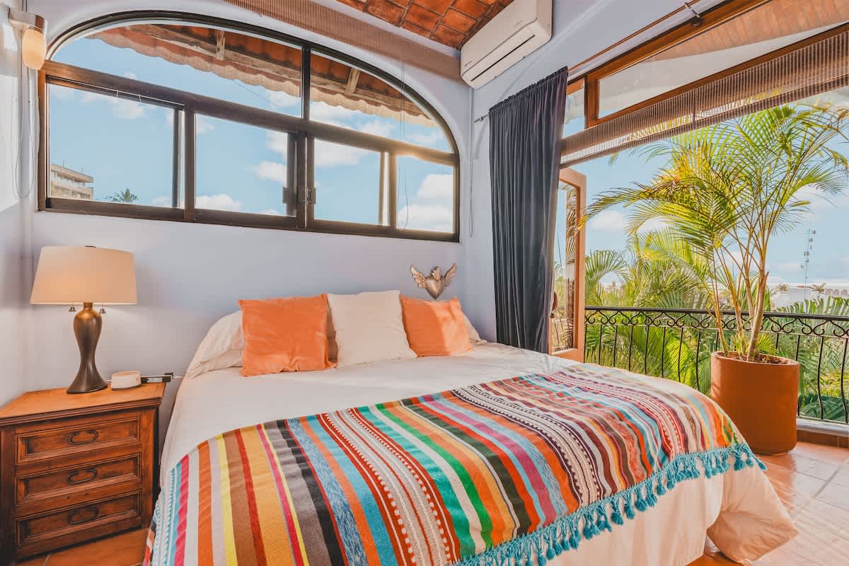 Bright bedroom with a large bed adorned with a colorful striped blanket and orange pillows, featuring an arched window overlooking a balcony with lush greenery and an air conditioning unit.