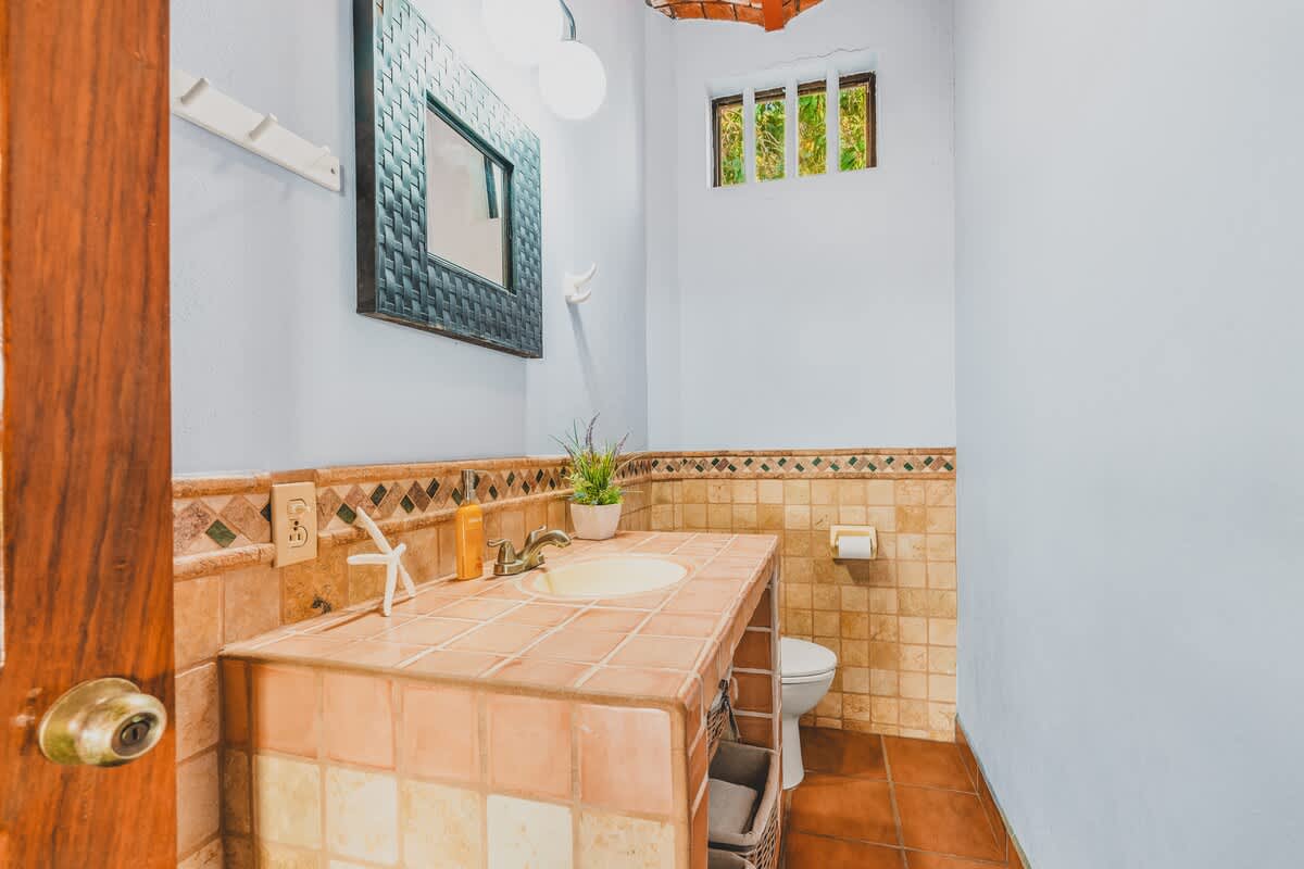 A compact bathroom featuring a tiled vanity with an integrated sink, a dark framed mirror, a toilet, and a small arched window. The walls are light blue with a decorative tiled wainscoting.