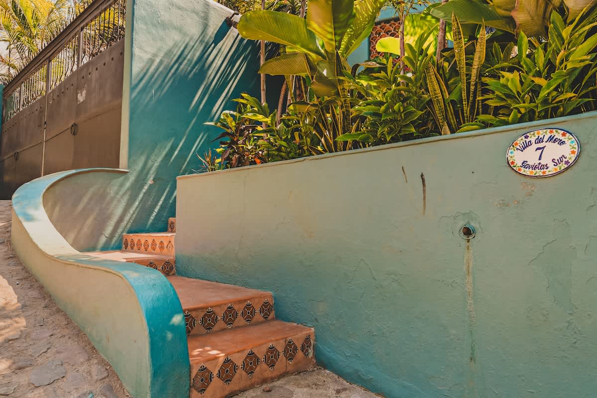 Terracotta-tiled steps with decorative patterns ascend alongside a curved, teal-colored wall, leading up to the property entrance. Lush green plants and a house number plaque are visible, with a dark metal gate in the background.
