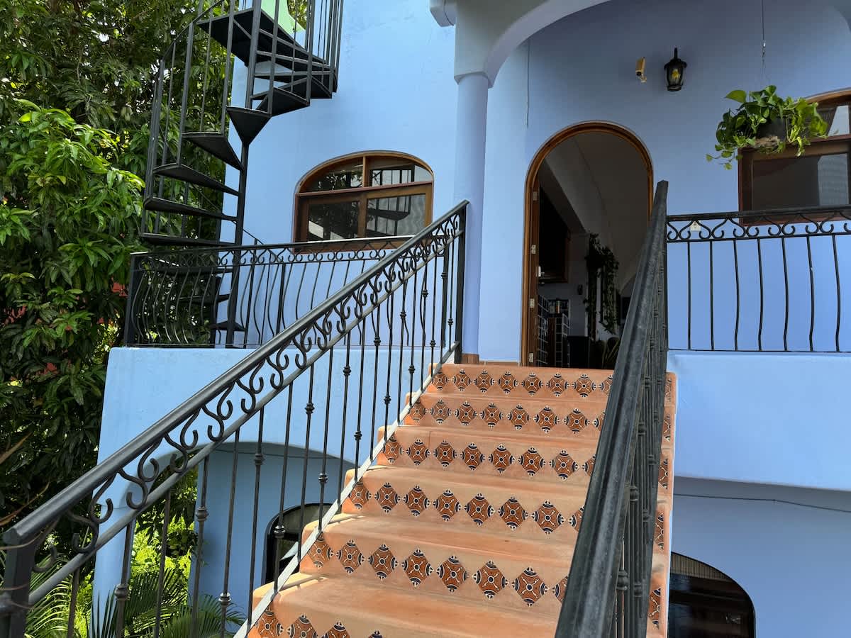 The exterior entrance of a light blue building, featuring a grand staircase with decorative terracotta tiles and a black wrought iron railing. A black spiral staircase is also visible to the left, leading to upper levels.