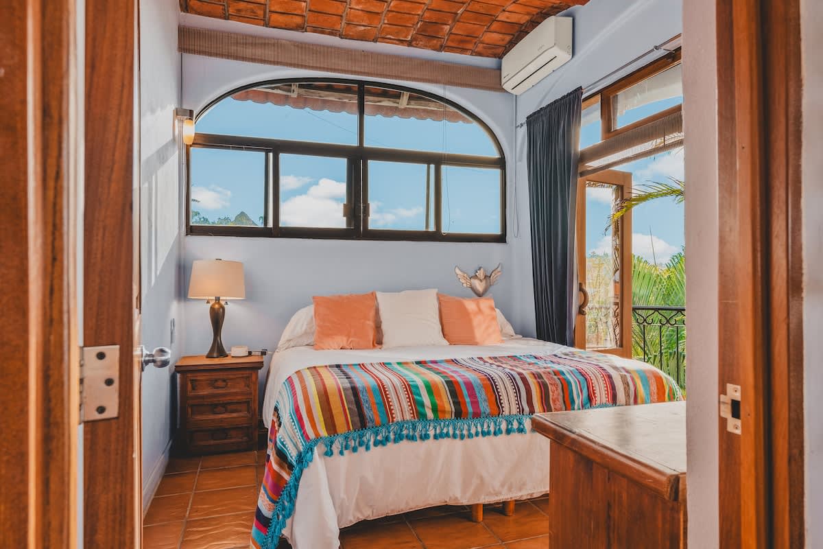 A cozy bedroom with a queen-sized bed adorned with a colorful striped blanket, a wooden nightstand with a lamp, and an arched window and glass door leading to a small balcony. The ceiling features exposed terracotta tiles.