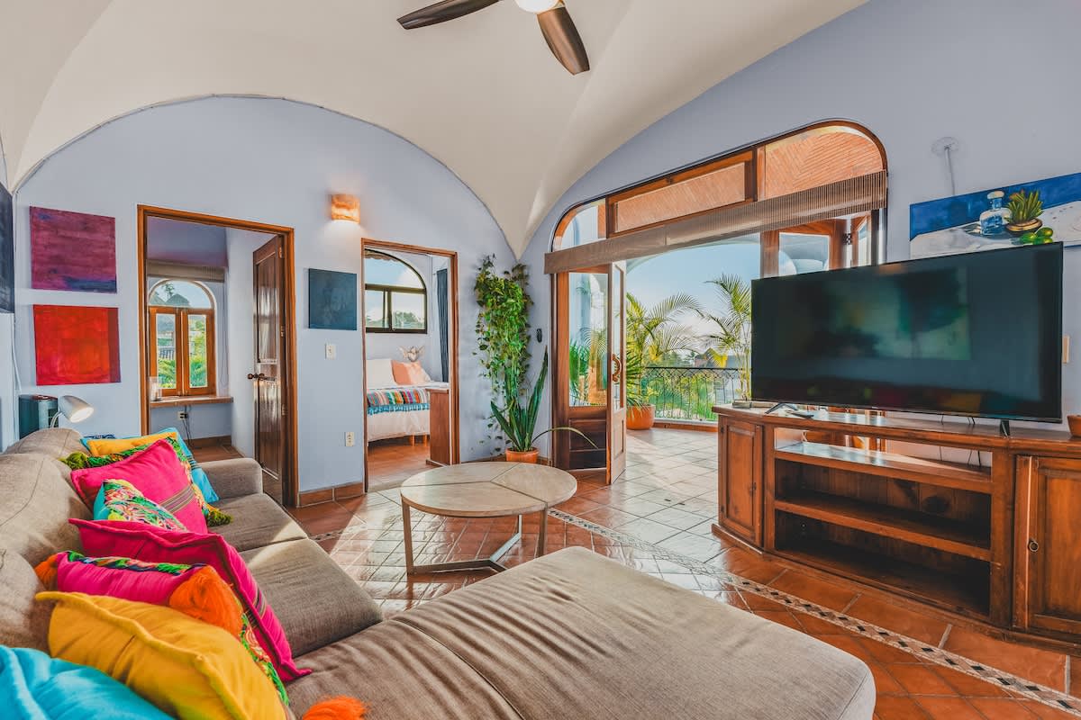 Living room with a large flat-screen TV on a wooden stand, a sectional sofa, and double doors opening to a balcony with tropical foliage and distant views.