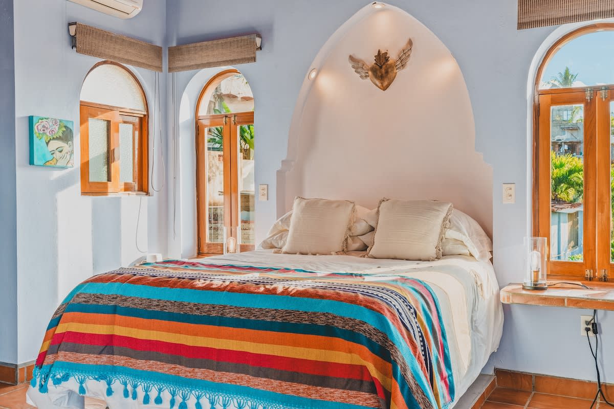 A sunlit bedroom showcasing a queen bed with a colorful striped blanket, an arched headboard, and two arched windows with wooden frames. An air conditioning unit is visible above one window.
