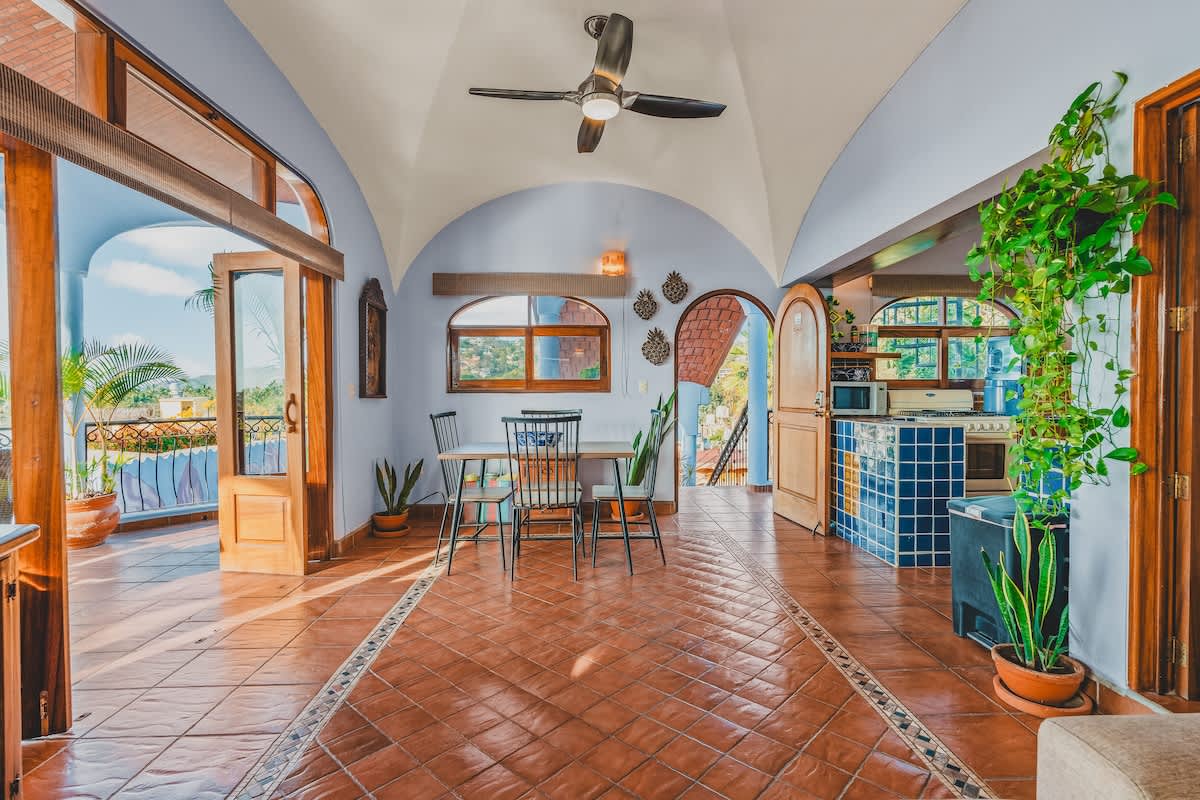 An open-concept living space features a central dining table, terracotta tile floors with decorative inlays, and a vaulted ceiling with a fan. Large glass doors open to a balcony with outdoor views, while a kitchen with blue tiled counters is visible to the right.