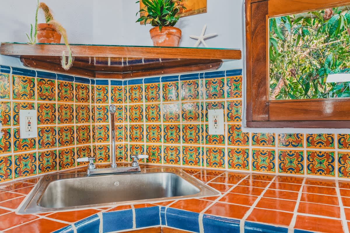 A detailed view of a kitchen corner featuring a stainless steel sink, chrome faucet, and a vibrant, multi-colored tiled backsplash. The countertop is a mix of blue and orange tiles, with a small wooden-framed window above.