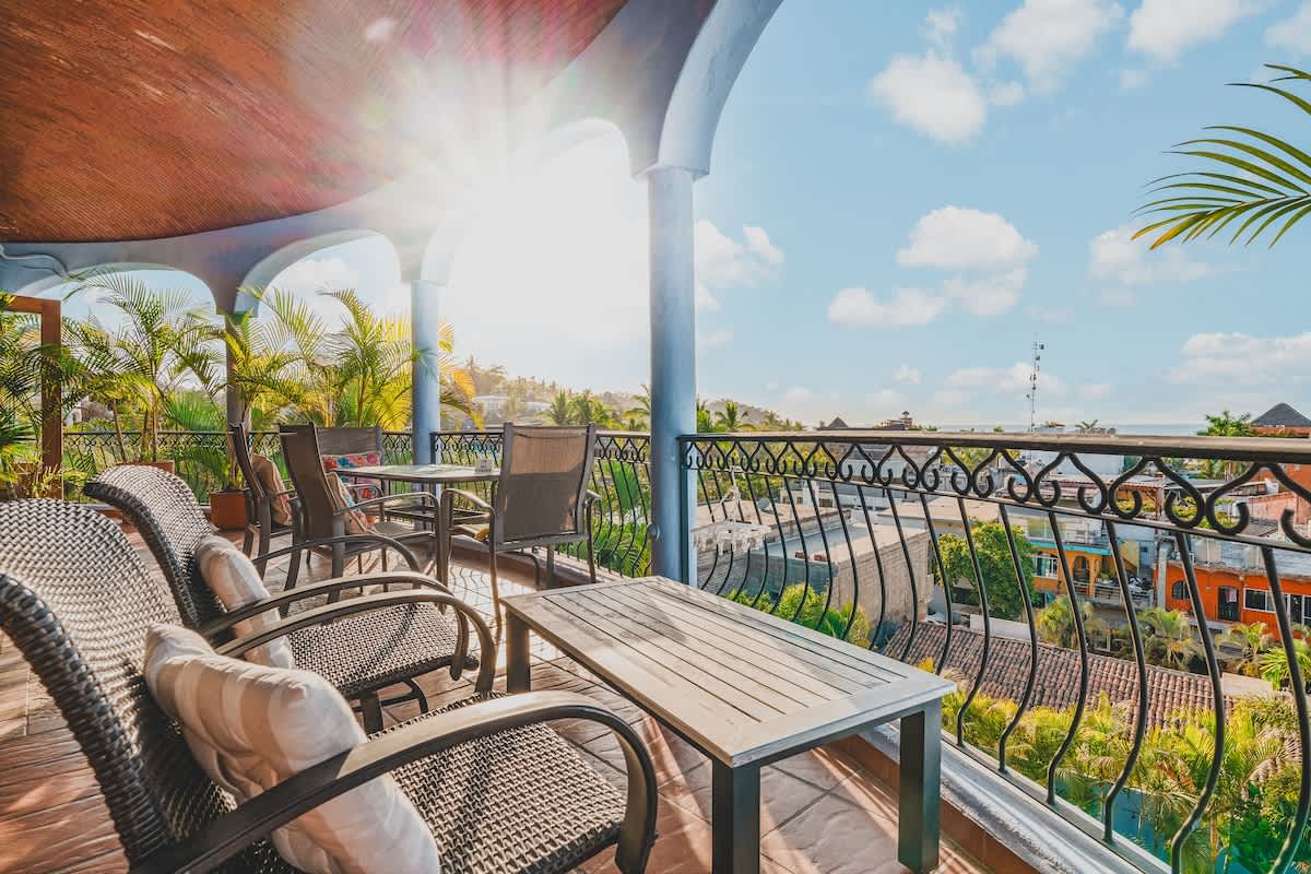A sun-drenched balcony with comfortable outdoor seating, including a wooden bench and wicker chairs. The view extends over the colorful town rooftops to the distant ocean under a bright blue sky.