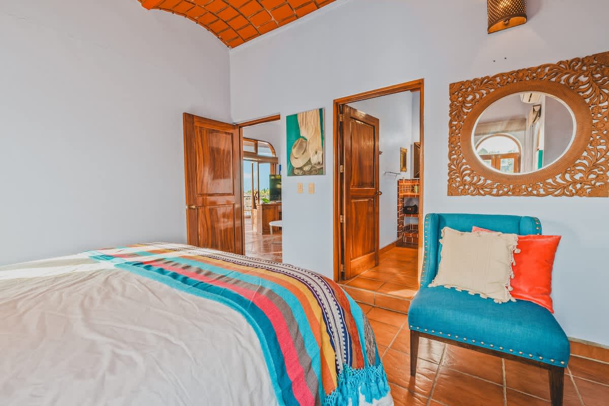 A bedroom with a queen bed featuring a colorful striped blanket, a decorative carved wooden mirror, and two wooden doors, one leading to the main living area and the other to an en-suite bathroom.