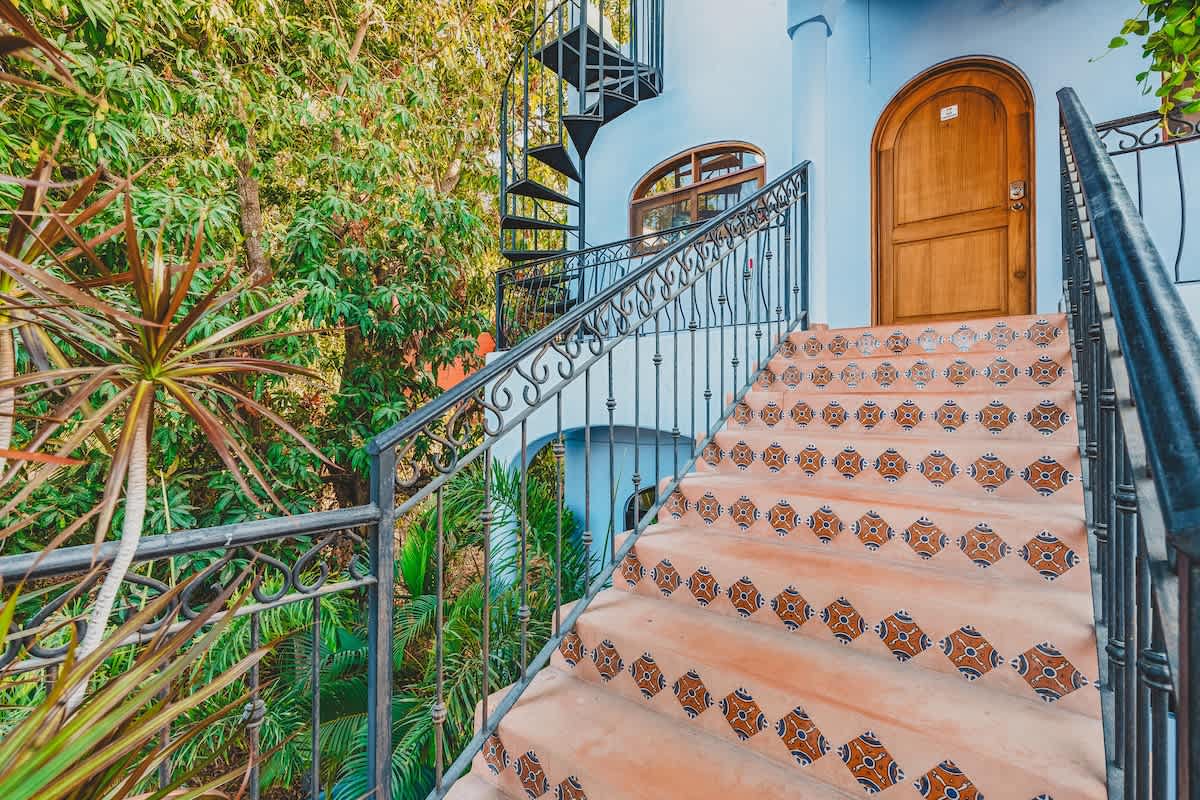An inviting outdoor staircase with decorative patterned tiles and a black wrought-iron railing, leading up to a light blue building with an arched wooden door, surrounded by lush tropical foliage. A spiral staircase is visible in the background.