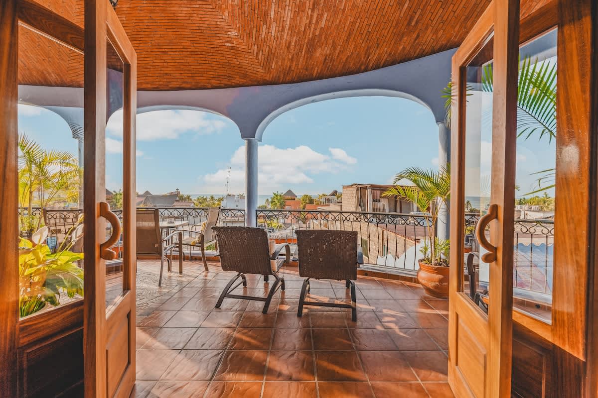 A welcoming view from indoors, looking out through open wooden double doors onto a spacious balcony with terracotta-tiled flooring. The balcony features outdoor seating and offers a bright view of the sky and surrounding rooftops.