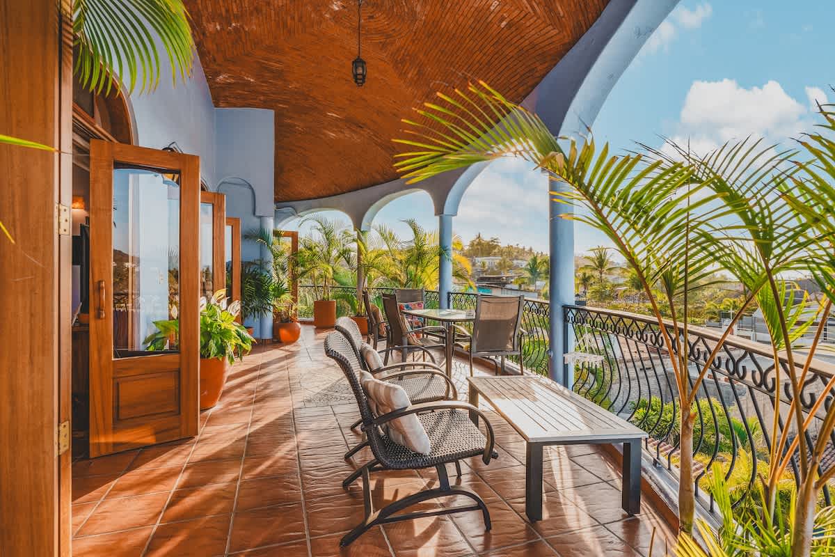 A wide-angle view of the spacious, covered balcony, showcasing various outdoor seating options and potted plants. The terracotta-tiled floor extends along the length, offering an elevated perspective of the surrounding landscape.