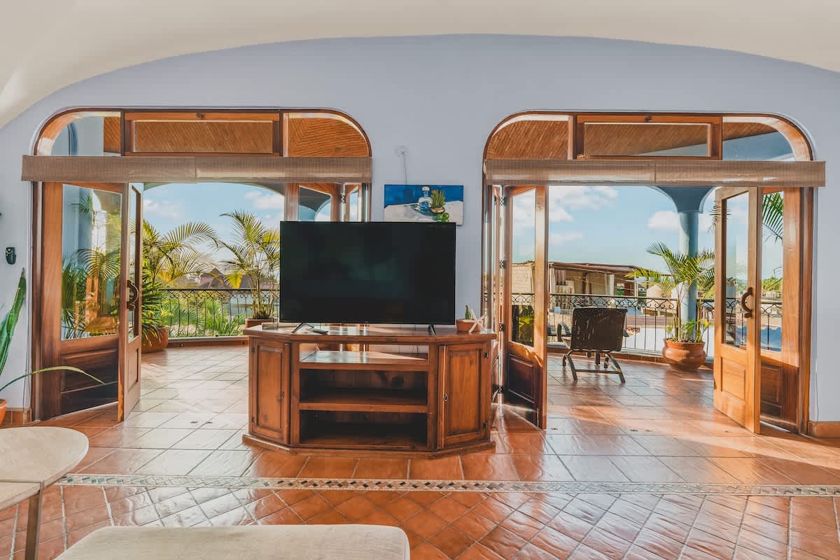 A bright living space with warm terracotta tiled floors, a large flat-screen TV on a wooden console, and two arched glass doors leading to an expansive outdoor balcony with lush palm trees.