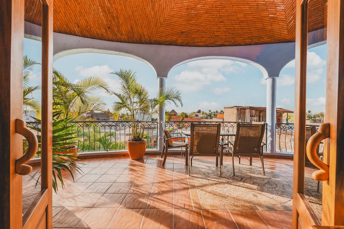 An expansive outdoor balcony with terracotta-tiled flooring and a curved, vaulted ceiling, furnished with a glass-top table and chairs. Lush palm trees frame the view of the sky and distant buildings.