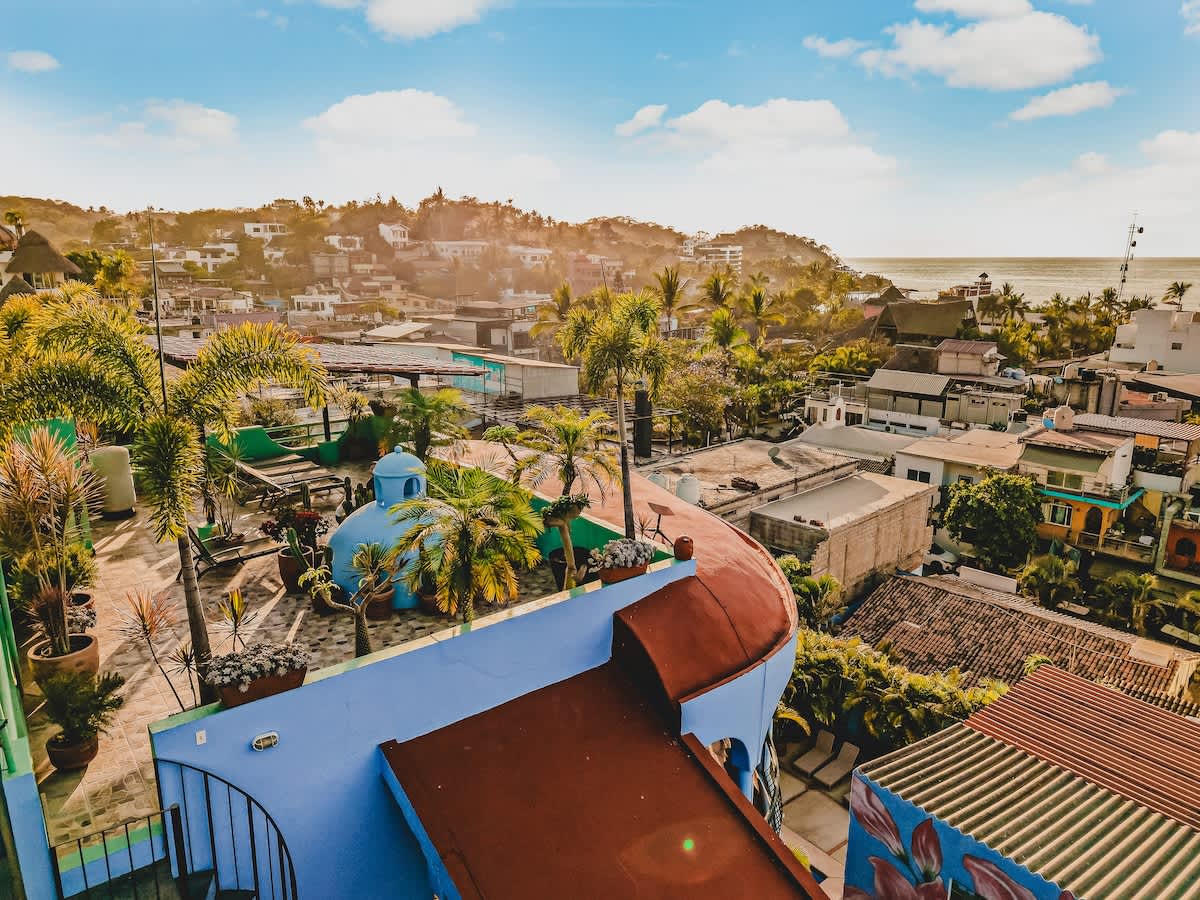 An aerial view captures the property's distinctive rooftop with a light blue dome and lush palm trees, overlooking a vibrant coastal town nestled against rolling hills with the sparkling ocean in the distance.