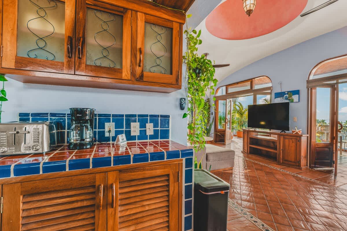 A section of the kitchen counter with blue tiled backsplash, a coffee maker, and a toaster, looking into an adjacent living area with a television and an open doorway leading to a spacious balcony.