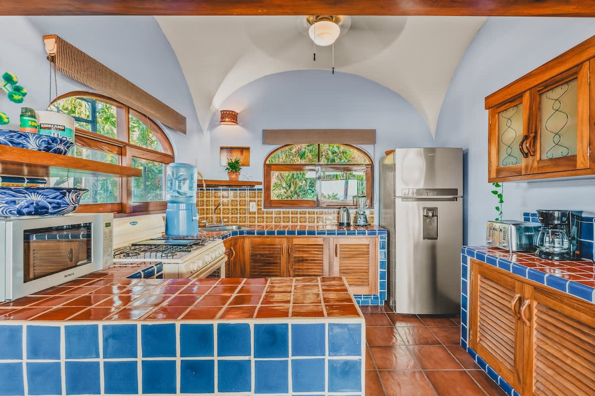 Colorful kitchen with blue and orange tiled countertops and backsplash, wooden cabinetry, a gas stove, microwave, and a stainless steel refrigerator, all under an arched ceiling.