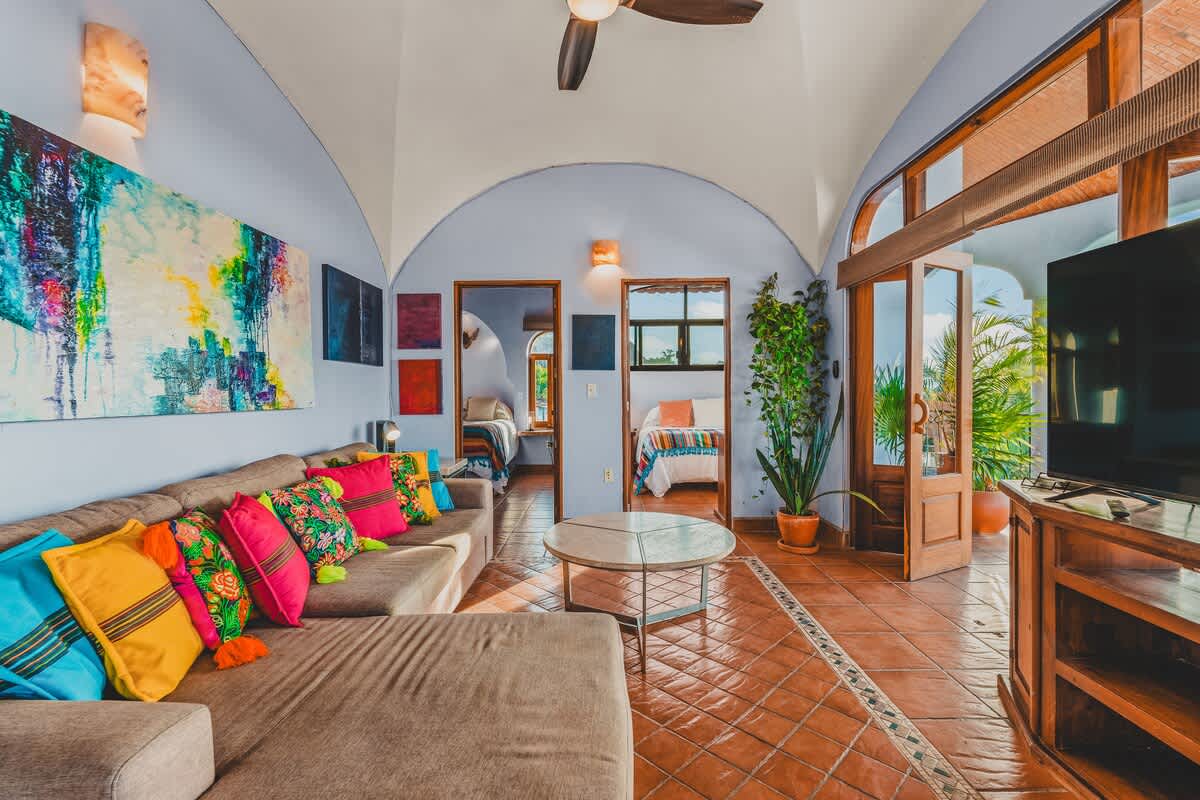 Wide view of the living room featuring a sectional sofa with vibrant pillows, a round coffee table, and a large flat-screen TV, with views into two adjacent bedrooms and a balcony.