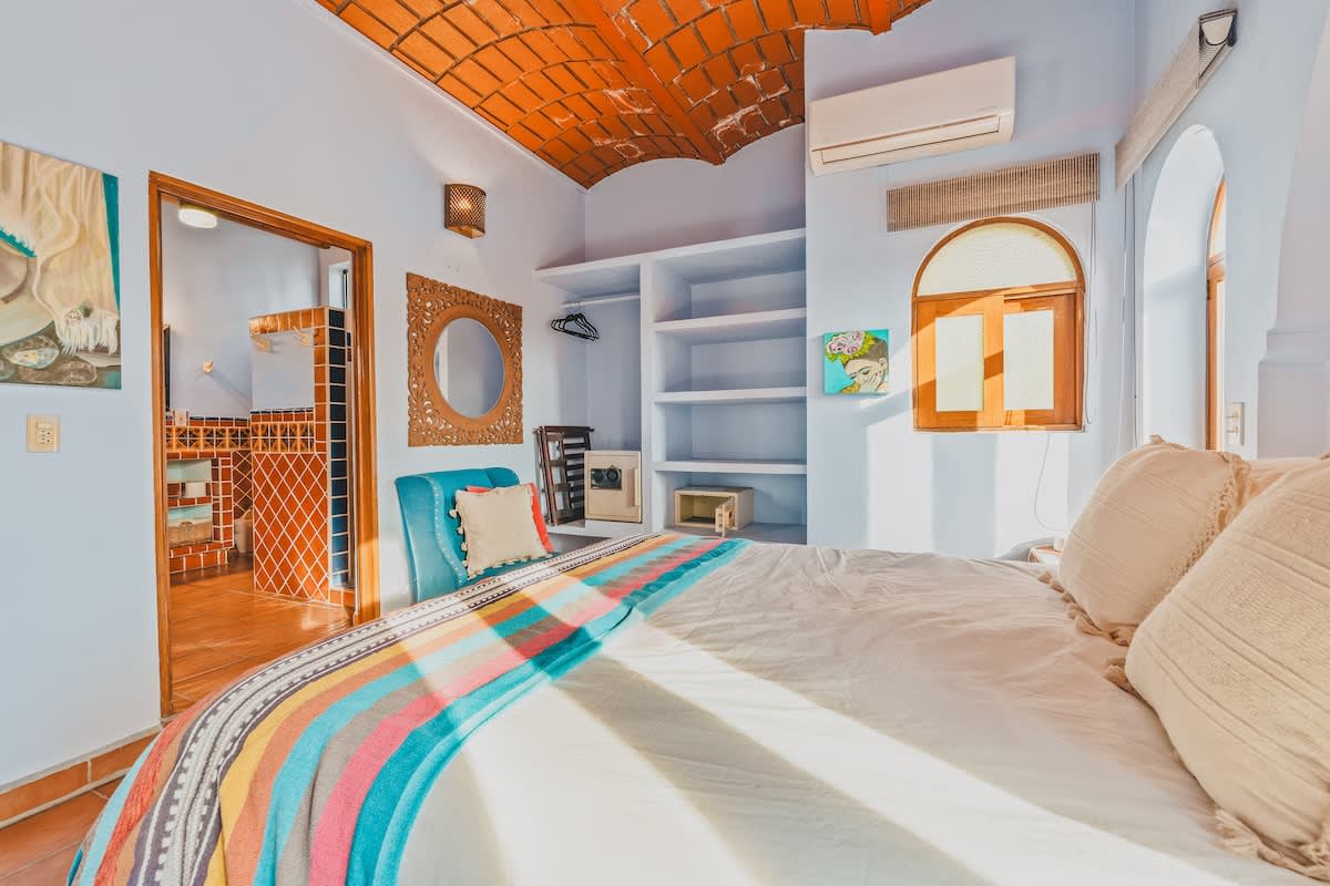 A bright bedroom featuring a queen bed with a colorful blanket, built-in open shelving, a decorative mirror, and a blue accent chair. An open doorway leads to an en-suite bathroom, and the ceiling has exposed terracotta tiles.