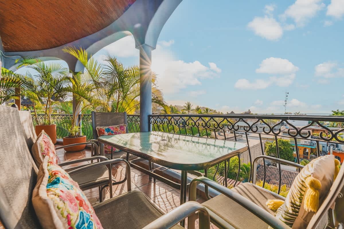 A sunny outdoor balcony featuring a glass-top dining table with four chairs, surrounded by a decorative wrought-iron railing, potted palm trees, and a distant view of the ocean and town.