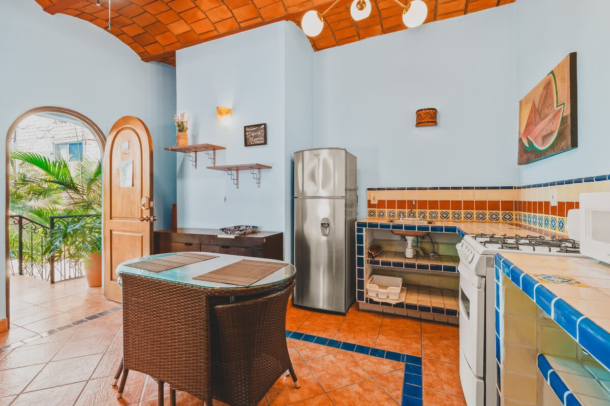 A vibrant kitchen features a stainless steel refrigerator, a white stove, and tiled countertops with colorful accents. A wicker dining table sits in the center, under a terracotta vaulted ceiling.
